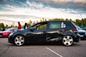 Black car with visible side damage parked in a lot, people in background.