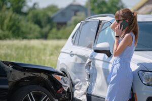 Woman on phone next to damaged car after an accident, seeking assistance.