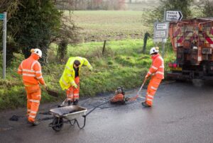 Road workers in orange safety gear repairing a pothole on a rural road with equipment.