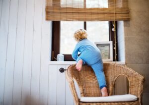 Toddler in pajamas climbing on a wicker chair near an open window, illustrating a fall risk.