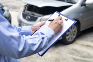 Insurance agent writing on clipboard in front of a damaged car, assessing vehicle damage for an insurance claim.