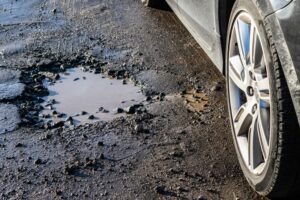 A close-up of a car tire next to a large, water-filled pothole on a damaged road.
