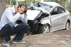 Man distressed by a car accident, sitting beside a wrecked vehicle with his head in his hands.