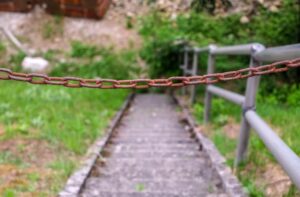 Rusty chain blocking access to a steep, overgrown staircase in an outdoor setting.