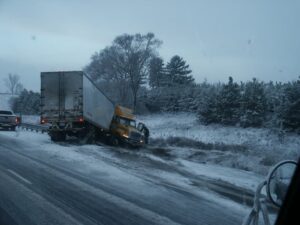 A jackknifed truck on a snowy roadside, with another vehicle and trees in the background.