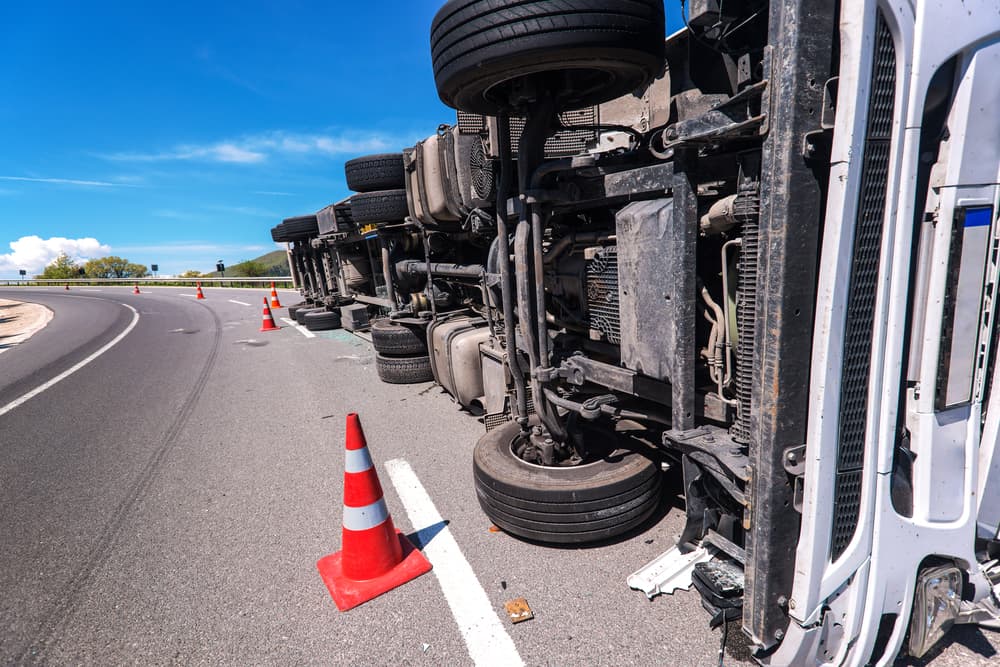 Overturned truck on a highway with traffic cones around, showing exposed underside and wheels.