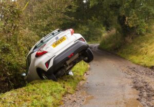 A white car partially overturned in a ditch beside a narrow, muddy country road.