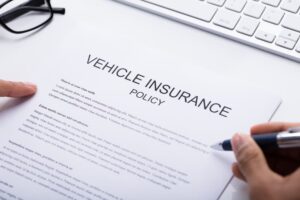 A person reviewing a vehicle insurance policy document with a pen, near a keyboard and glasses.