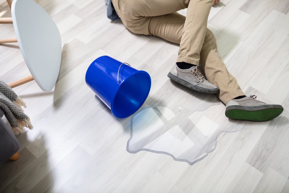 Person on the floor after slipping on water spilled from an overturned blue bucket.