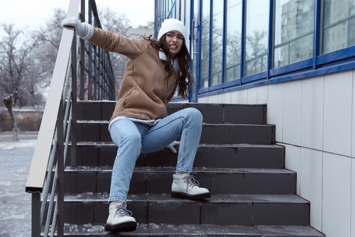 Person slipping on icy outdoor stairs, wearing a beige jacket, blue jeans, and winter boots, trying to grab the railing for support on a cold day with snow visible on the ground.